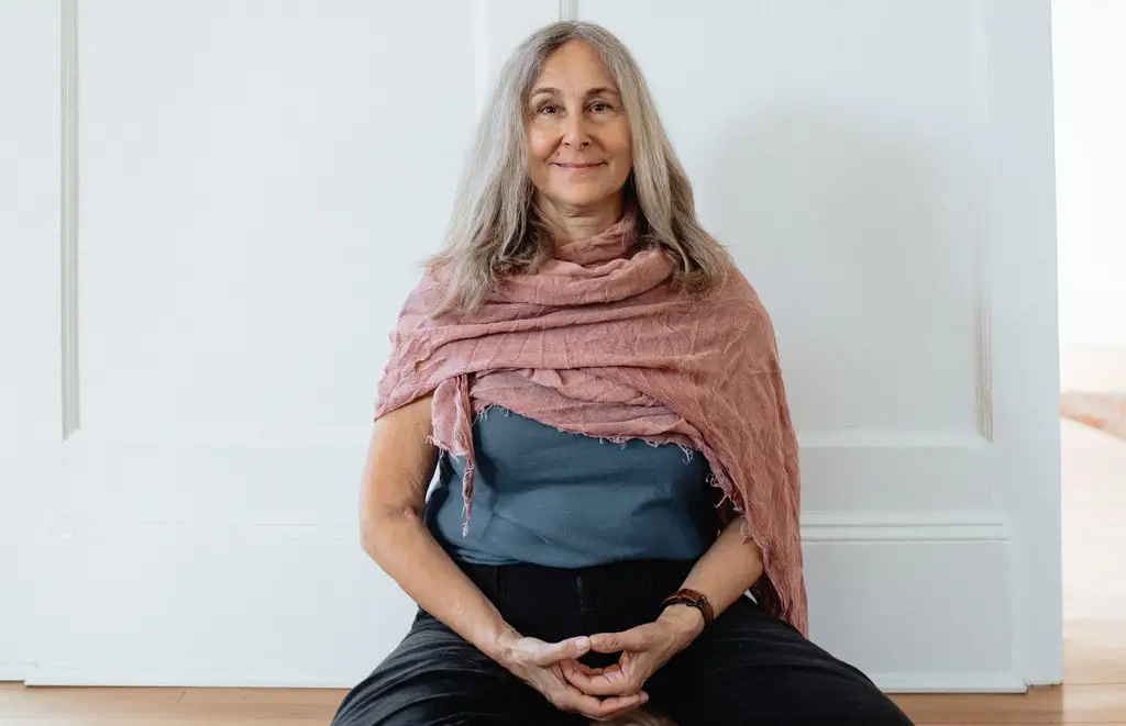 Older woman with long gray hair sits on the floor against a light wall, wearing a pink shawl and smiling gently.