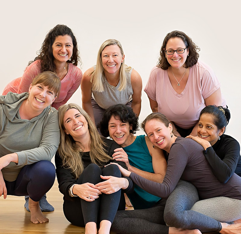 Group of women in yoga attire smiling together on a studio floor against a light wall, with open space on the left.
