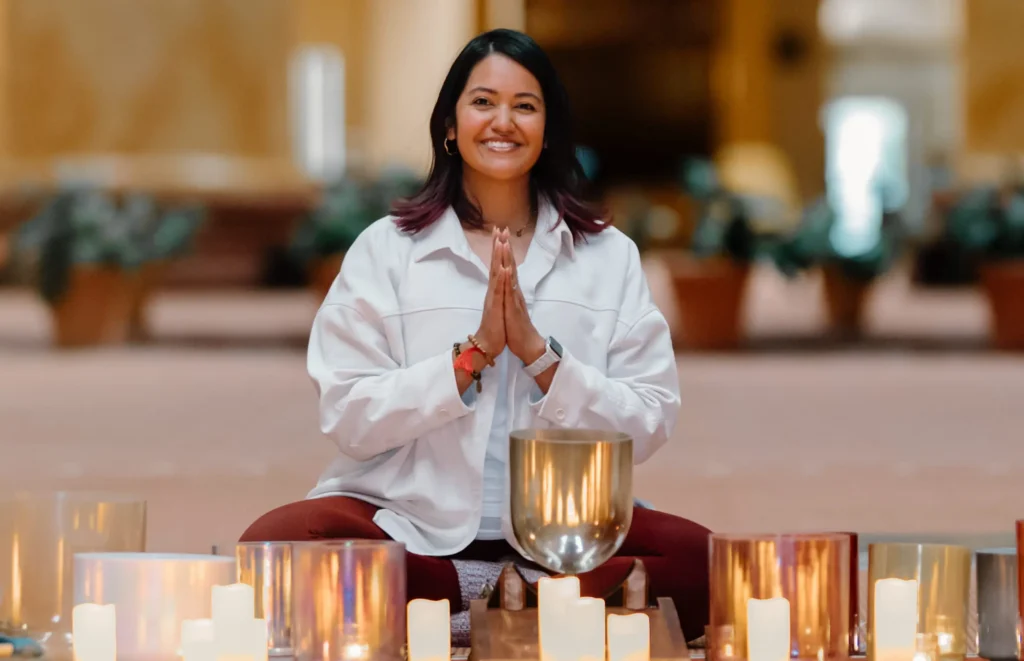 Woman sits cross-legged with hands in prayer, smiling, surrounded by lit candles and metal singing bowls in a softly lit room.