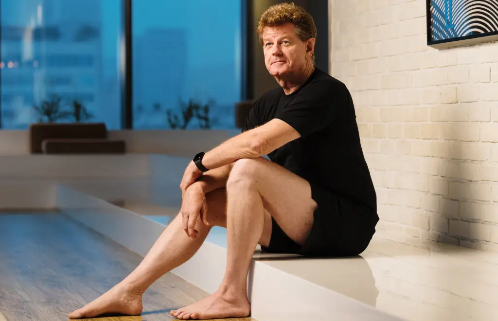 Middle-aged man in black athletic wear sits barefoot on a studio ledge beside a white brick wall, looking off to the side.