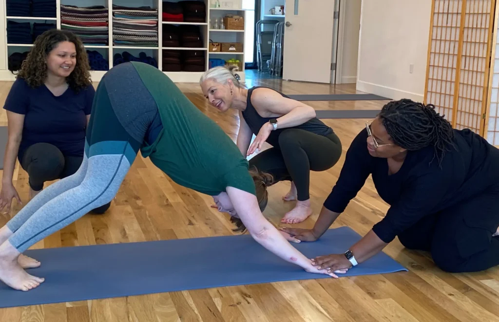 Yoga instructor crouches to guide a student in downward dog while two other students watch in a bright studio with mats and shelves.